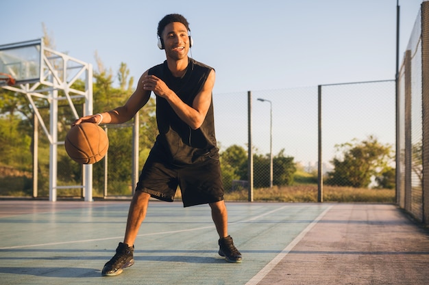 Joven sonriente feliz haciendo deporte, jugando baloncesto al amanecer, escuchando música en auriculares