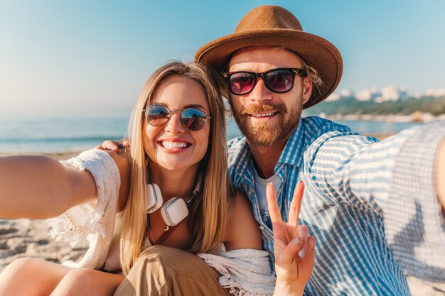 Joven sonriente feliz hombre y mujer en gafas de sol sentado en la playa de arena tomando fotos selfie en la cámara del teléfono