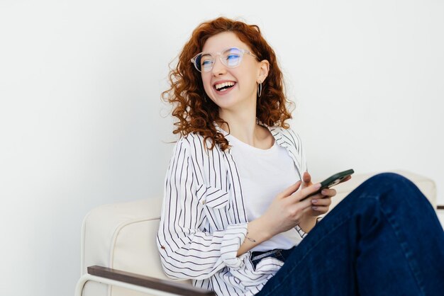 Una joven sonriente y feliz con el pelo rojo enviando mensajes de texto por teléfono tirada en el sofá de casa