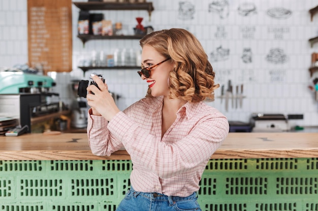 Foto gratuita joven sonriente con gafas de sol sentada en el mostrador del bar y tomando fotos con su pequeña cámara mientras pasa tiempo en el café