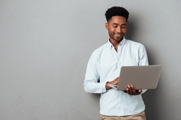 Joven sonriente hombre africano de pie y usando laptop