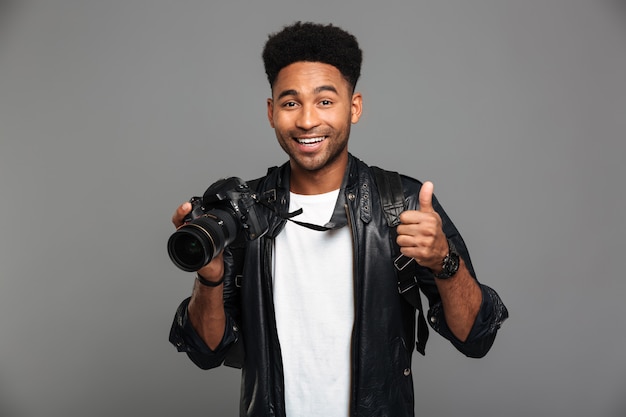 Joven sonriente hombre afroamericano con cámaras y mostrando el pulgar hacia arriba gesto, mirando
