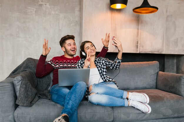 Joven sonriente hombre y mujer sentados en casa en invierno, sosteniendo la computadora portátil, escuchando auriculares, pareja en el tiempo libre juntos, haciendo una foto selfie en la cámara del teléfono inteligente, feliz, positivo, citas