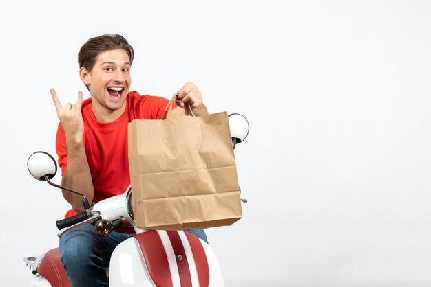 Joven sonriente loco mensajero emocional en uniforme rojo sentado en scooter sosteniendo una bolsa de papel en la pared blanca