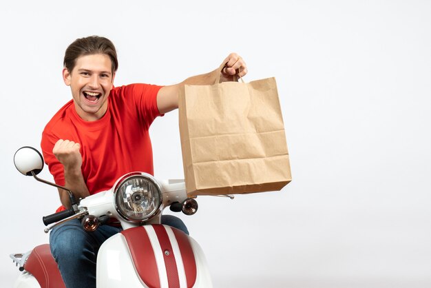 Joven sonriente mensajero confiado en uniforme rojo sentado en scooter sosteniendo una bolsa de papel en la pared blanca