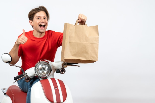 Joven sonriente mensajero en uniforme rojo sentado en scooter sosteniendo una bolsa de papel y haciendo un gesto de ok en la pared amarilla