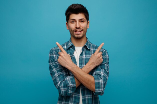 Joven sonriente mirando a la cámara manteniendo las manos cruzadas apuntando a lados aislados en fondo azul con espacio para copiar