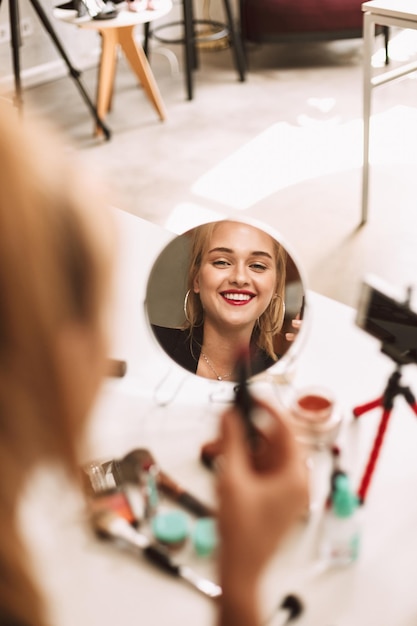 Foto gratuita joven sonriente mirando felizmente en un pequeño espejo en la mesa con lápiz labial en la mano mientras graba un nuevo video para vlog