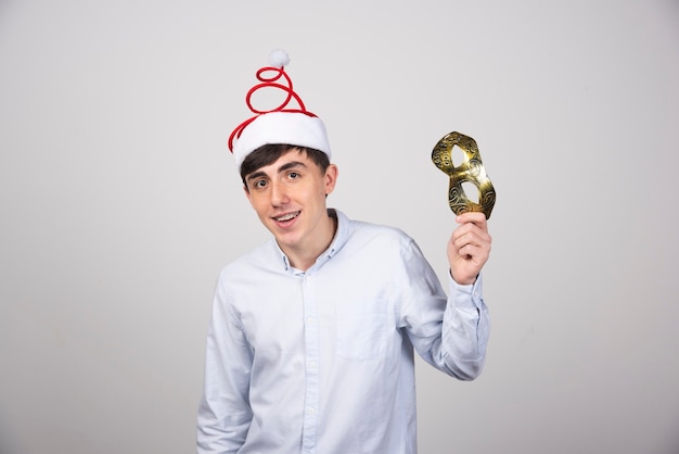 Joven sonriente modelo de hombre de pie en el sombrero de Navidad con máscara de carnaval
