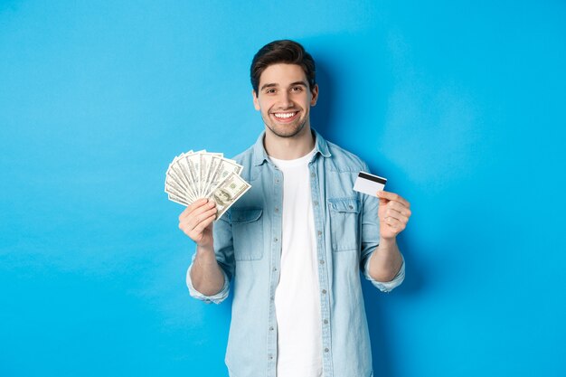 Joven sonriente mostrando dólares en efectivo y tarjeta de crédito, de pie sobre fondo azul.