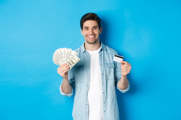 Joven sonriente mostrando dólares en efectivo y tarjeta de crédito, de pie sobre fondo azul.