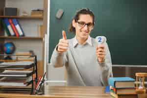 Foto gratuita joven sonriente mostrando los pulgares hacia arriba profesor masculino con gafas sentado en el escritorio sosteniendo números de matemáticas con herramientas escolares en el aula