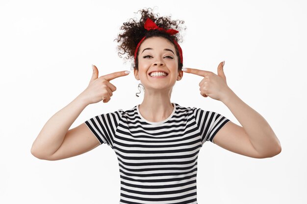 Una joven sonriente mostrando sus dientes blancos y perfectos, señalando la boca y luciendo feliz, parada en una camiseta contra fondo blanco