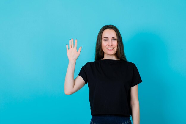 Una joven sonriente muestra un gesto de saludo levantando la mano sobre fondo azul.
