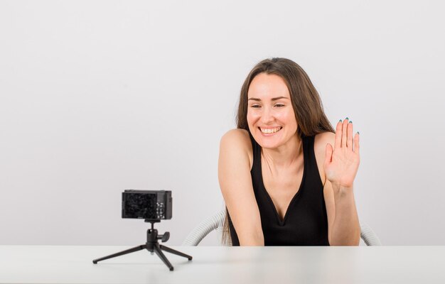 Una joven sonriente muestra un gesto de saludo a su pequeña cámara con fondo blanco