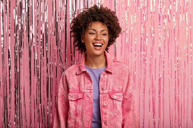 Joven sonriente mujer afroamericana tiene maquillaje mínimo, viste chaqueta rosa