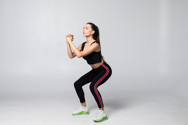 Joven sonriente mujer deporte haciendo sentadillas en la pared gris