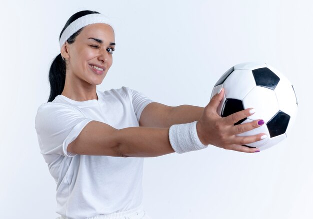 Joven sonriente mujer deportiva caucásica con diadema y muñequeras parpadea sosteniendo la bola aislada en el espacio en blanco con espacio de copia