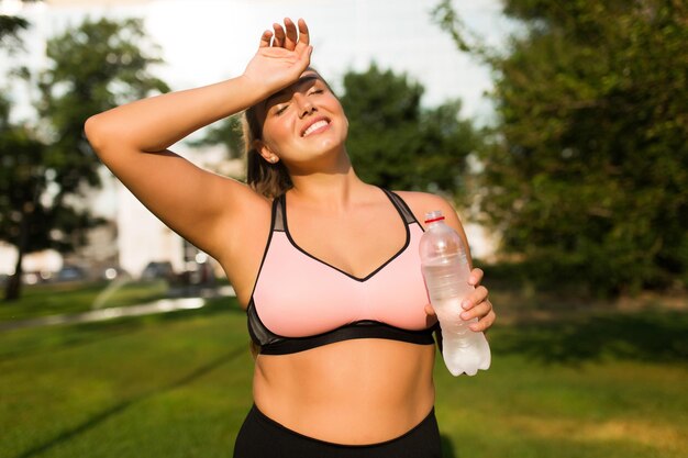 Joven y sonriente mujer de talla grande con top deportivo rosa sosteniendo una botella de agua pura en la mano cerrando los ojos soñadoramente mientras pasa tiempo en el parque de la ciudad