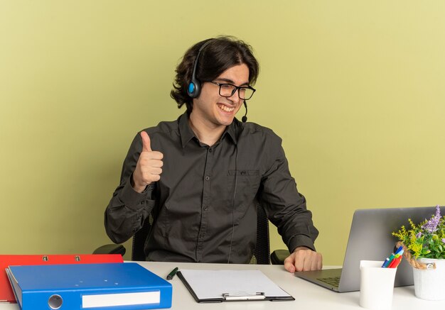 Joven sonriente oficinista hombre en auriculares con gafas ópticas se sienta en el escritorio con herramientas de oficina usando y mirando portátil Thumbs up