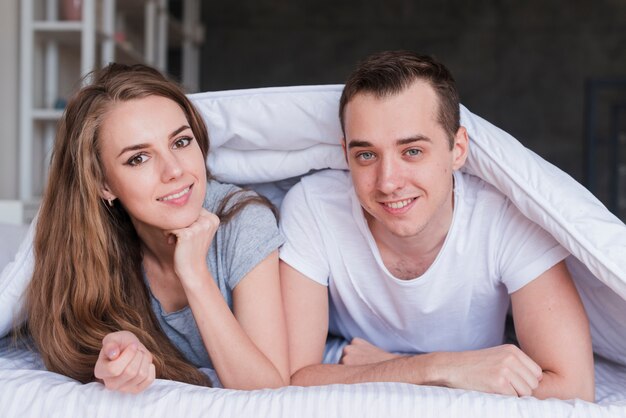 Joven sonriente pareja acostada en la cama bajo edredón