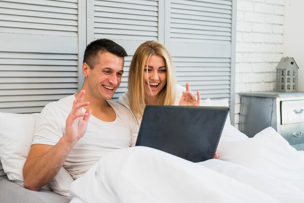 Joven sonriente pareja con portátil y manta en cama