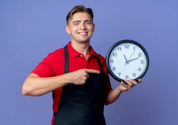 Joven sonriente peluquero masculino rubio en uniforme sostiene y apunta al reloj aislado en el espacio violeta con espacio de copia