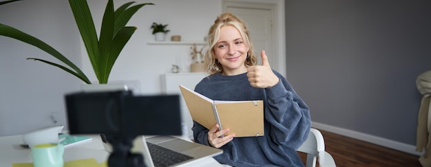 Foto gratuita una joven sonriente y positiva muestra los pulgares hacia arriba se sienta en una habitación con cámara digital y registros de portátil