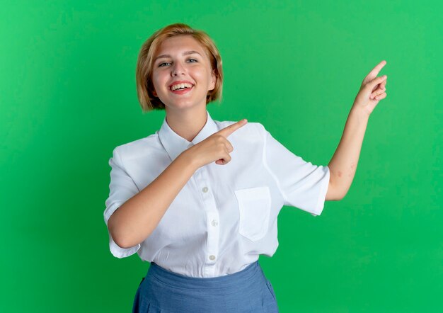 Joven sonriente rubia rusa apunta al lado aislado sobre fondo verde con espacio de copia