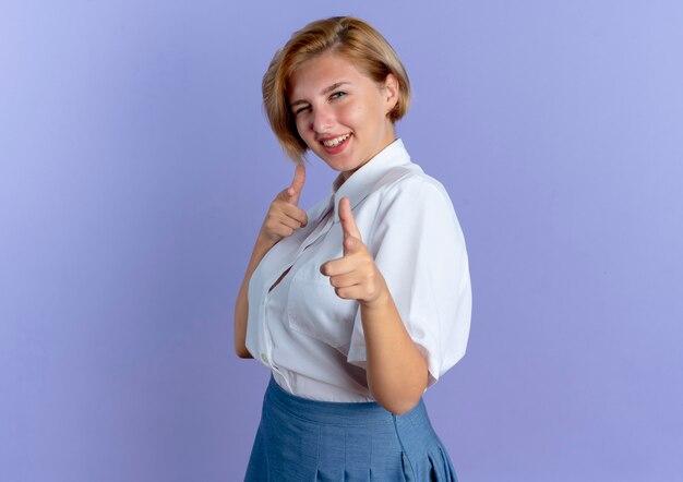 Joven sonriente rubia rusa está parado hacia los lados parpadea el ojo apuntando a la cámara aislada sobre fondo púrpura con espacio de copia