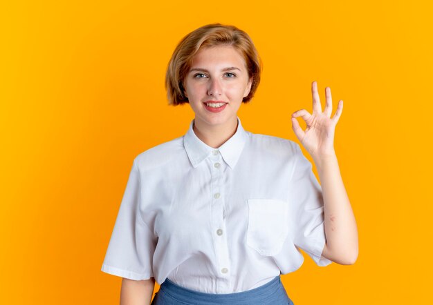 Joven sonriente rubia rusa gestos ok signo de mano aislado sobre fondo naranja con espacio de copia