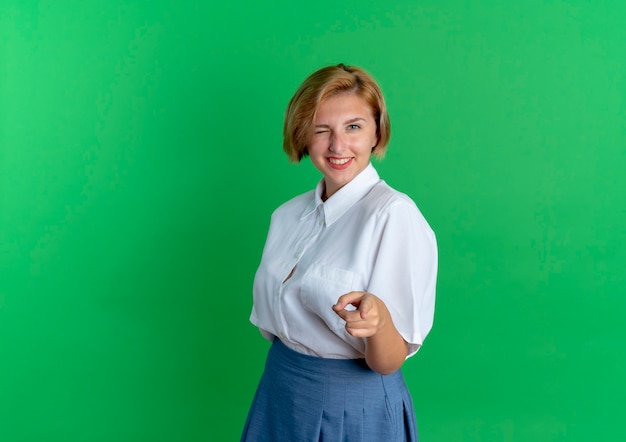 Joven sonriente rubia rusa parpadea y apunta a la cámara aislada sobre fondo verde con espacio de copia