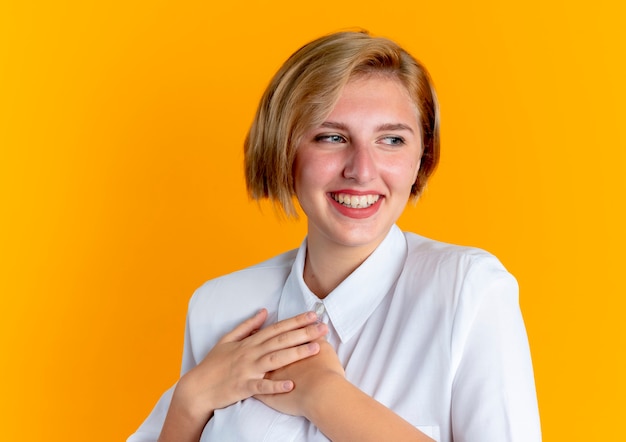 Joven sonriente rubia rusa pone las manos en el pecho mirando al lado aislado sobre fondo naranja con espacio de copia