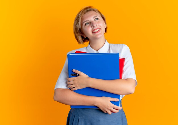 Joven sonriente rubia rusa sostiene carpetas de archivos mirando hacia arriba aislado sobre fondo naranja con espacio de copia