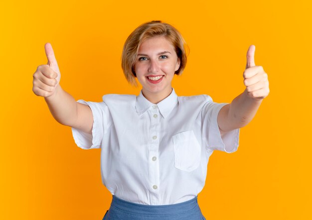 Joven sonriente rubia rusa Thumbs up con dos manos aisladas sobre fondo naranja con espacio de copia