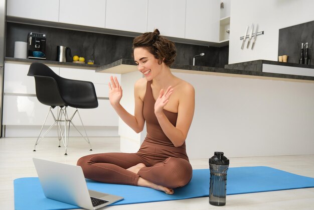 Una joven sonriente se sienta en una alfombrilla de goma deportiva con ropa activa y dice hola en un video chat en línea yoga cla