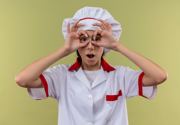 Joven sorprendida cocinera caucásica en uniforme de chef mira a través de los dedos aislados sobre fondo verde con espacio de copia