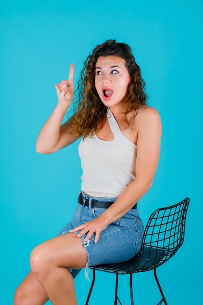 Una joven sorprendida está apuntando hacia arriba con el dedo índice sentándose en una silla de fondo azul