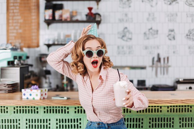 Joven sorprendida con gafas de sol y gorra de cumpleaños sentada en el mostrador del bar con batido en la mano y mirando asombrosamente a un lado en el café