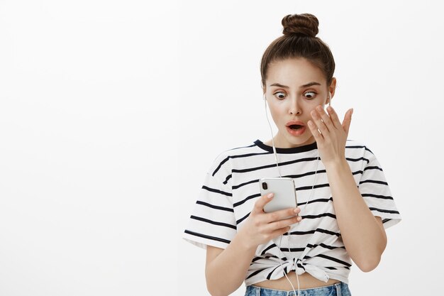 Joven sorprendida y preocupada jadeando mientras mira la pantalla del teléfono móvil, usando audífonos