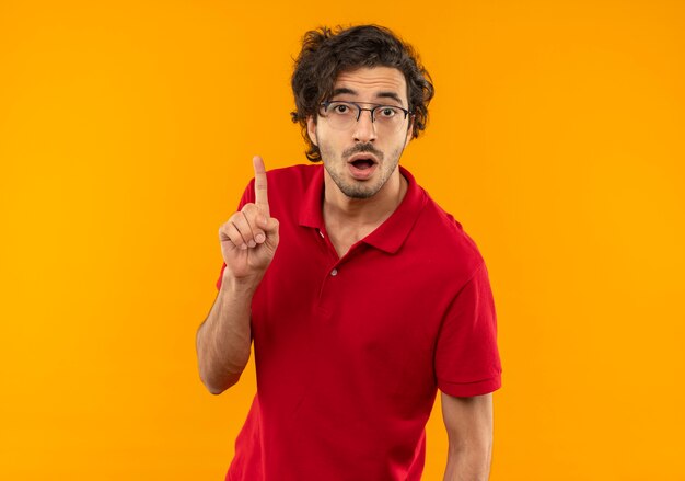 Joven sorprendido en camisa roja con gafas ópticas apunta hacia arriba y parece aislado en la pared naranja