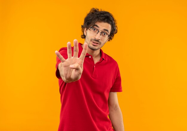 Joven sorprendido en camisa roja con gafas ópticas gestos signo de cuatro manos aislado en la pared naranja