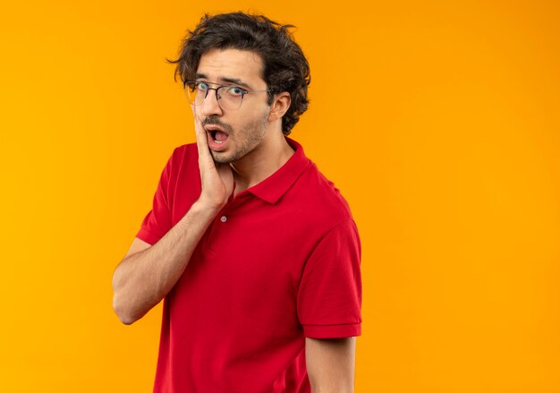 Joven sorprendido en camisa roja con gafas ópticas pone la mano en la cara y parece aislado en la pared naranja