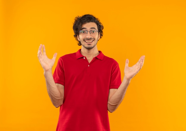 Joven sorprendido en camisa roja con gafas ópticas tiene las manos abiertas aisladas en la pared naranja