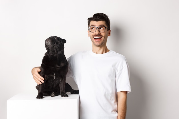 Foto gratuita joven sorprendido con gafas abrazando a su perro, dueño de la mascota y pug mirando la oferta promocional de la esquina superior izquierda, de pie sobre fondo blanco.