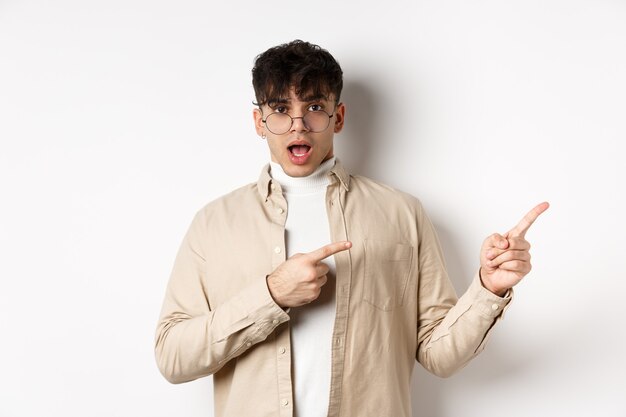 Joven sorprendido con gafas apuntando con el dedo a la derecha en el espacio vacío, preguntando por publicidad, de pie sobre fondo blanco.