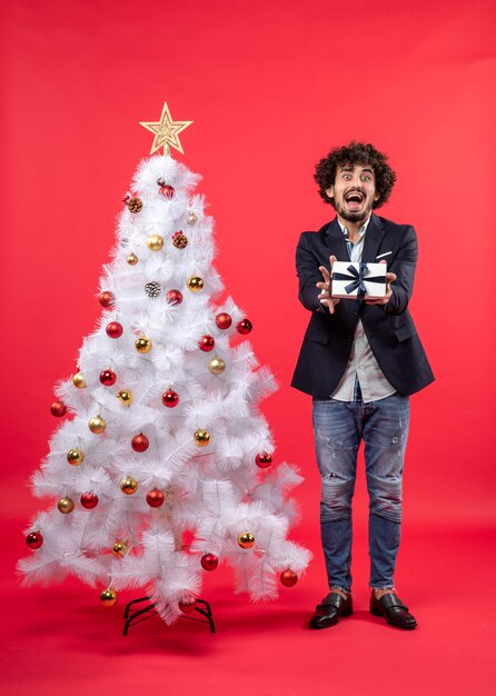 Joven sorprendido mostrando su regalo de pie cerca del árbol de Navidad blanco decorado en el lado derecho de rojo
