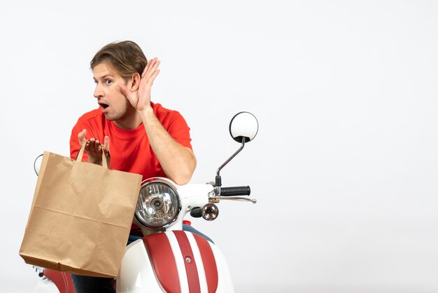 Joven sorprendido repartidor en uniforme rojo sentado en scooter sosteniendo una bolsa de papel y escuchando los últimos chismes en la pared blanca