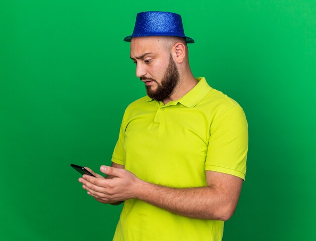 Joven sorprendido con sombrero de fiesta azul sosteniendo y mirando el teléfono