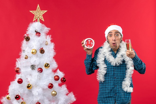 Joven sorprendido con sombrero de santa claus y sosteniendo una copa de vino y reloj de pie cerca del árbol de Navidad